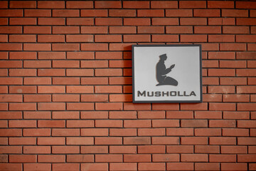 Musholla sign with praying silhouette on red brick wall, Islamic prayer room designation board.