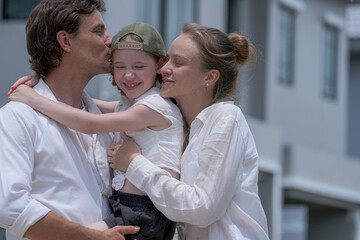 Fototapeta premium A happy Caucasian family, a father holding his daughter, and a mother, stand together outdoors. The man and child are smiling and pointing at something off-screen.