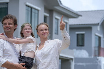 Obraz premium A hopeful young family looks towards their future outside a new modern home. The father holds their daughter, who points excitedly, as the mother smiles, dreaming of their life together.