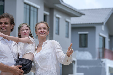 Obraz premium A hopeful young family looks towards their future outside a new modern home. The father holds their daughter, who points excitedly, as the mother smiles, dreaming of their life together.