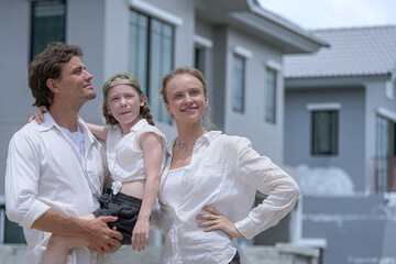 Obraz premium A hopeful young family looks towards their future outside a new modern home. The father holds their daughter, who points excitedly, as the mother smiles, dreaming of their life together.