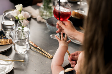 Close-up of a baby interacting with an adult at a dining table during a social gathering, with dessert, flowers, and a red drink creating a warm family celebration atmosphere.