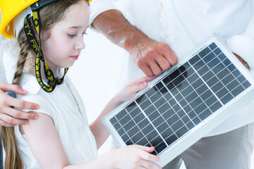 A curious young girl in a yellow hard hat learns about renewable energy. An adult teaches her about a solar panel, sparking interest in sustainability and future green technology.