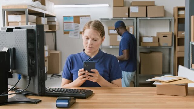 Medium shot of cheerful Caucasian female courier service associate picking up smartphone, calling client to inform on arrival of package, while working on computer in office