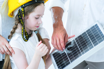 A curious young girl in a yellow hard hat learns about renewable energy. An adult teaches her about a solar panel, sparking interest in sustainability and future green technology.