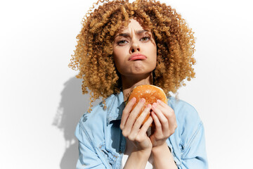 Young woman with curly hair holding a hamburger, showcasing a playful yet disappointed expression...