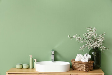 Vase with blooming twigs and bath supplies on chest of drawers near green wall in bathroom, closeup