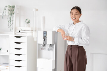 Young Asian businesswoman with glass of water near cooler in office