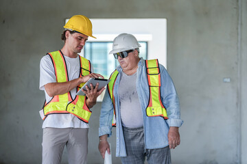 A young foreman shows a digital plan on a tablet to a senior architect holding blueprints. Two construction workers discuss a project at an unfinished building site.
