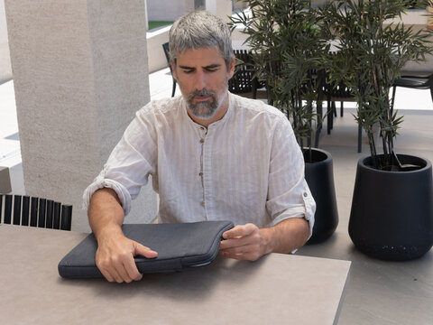 Man with gray hair unzipping laptop sleeve on a modern outdoor terrace, ready for remote work or studying
