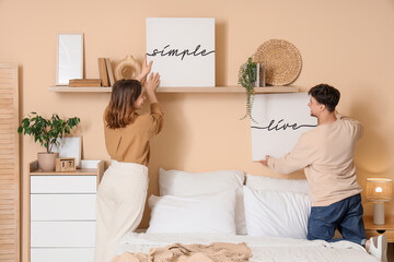 Young couple hanging posters on beige wall in bedroom, back view
