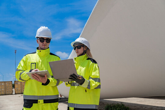 Two engineers in high visibility gear use a laptop to work in front of a large wind turbine blade at a construction site. the integration of technology in renewable energy and wind power projects.
