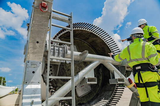 Two engineers work on a wind turbine component, with one using a laptop and the other inspecting parts on a ladder. renewable energy technology, wind power projects, and teamwork in the energy sector.