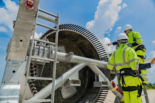 Two engineers work on a wind turbine component, with one using a laptop and the other inspecting parts on a ladder. renewable energy technology, wind power projects, and teamwork in the energy sector. - Powered by Adobe