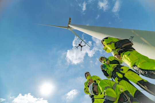 engineers conducting an aerial inspection of a wind turbine using a drone. The scene highlights modern technology’s role in renewable energy maintenance, showcasing collaboration and innovation. - Powered by Adobe