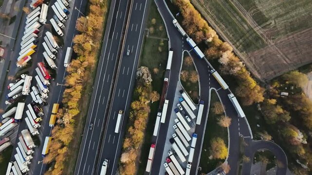 aerial view truck parking rows beside highway with long queue of trailers, autumn trees lining roadside, slow moving traffic, logistics staging and parked fleet, industrial depot atmosphere