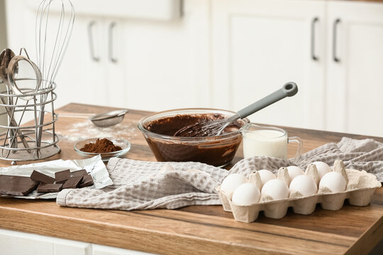 Bowl with raw dough for preparing tasty chocolate cake and baking utensils on wooden table in kitchen