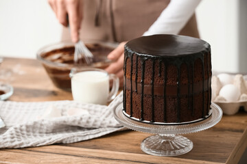 Young woman with ingredients and tasty chocolate cake on table in kitchen, closeup