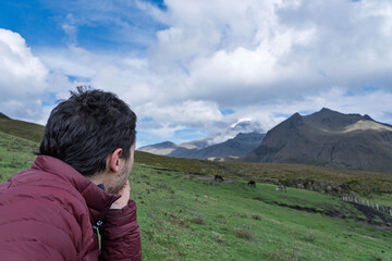 Man wearing a red down jacket, looking at a snow-capped mountain in the middle of a beautiful valley full of green hills.