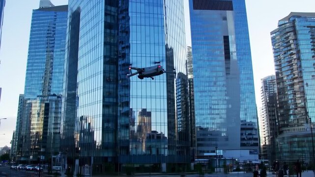 A drone flies near reflective glass skyscrapers under a bright blue sky