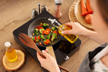 Woman frying vegetables with sunflower oil on mini stove in kitchen, closeup