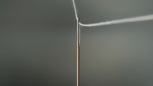 Macro view of a hand guiding a white thread through the eye of a sewing needle, steady motion and careful precision, craftsmanship and patience concept