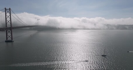 Cinematic aerial drone establishing shot towards the 25th April Bridge and waterfront in Lisbon, Portugal, Europe. Sunny and bright with clouds, fog, mist covering the bridge