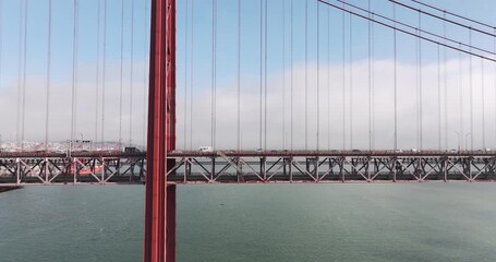 Aerial drone shot towards the 25th April Bridge in Lisbon, Portugal, Europe. Sunny and bright with clouds in the background. Cars driving on bridge, side view. Close-up perspective