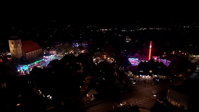 Night scene o of American village during festival funfair. Historic church tower in German town and lighting carnival fairground ride. Aerial wide shot.
