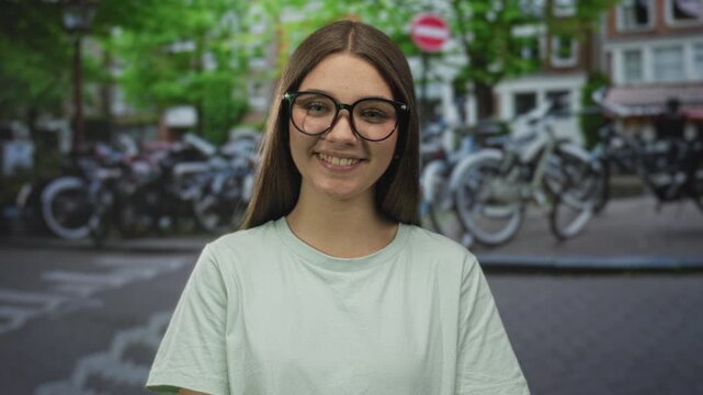 Girl with glasses smiles on a busy city street lined with bicycles wearing a light green shirt; youthful joy.