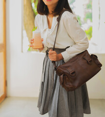 The body of a girl in a gray skirt and white shirt stands while holding a glass of cold drink, carrying a beautiful dark brown leather bag.