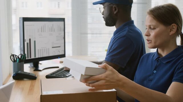 Medium shot of two multiethnic associates working at courier delivery company office, African American male manager filling out report on computer while female employee is carrying parcels