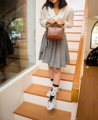 The body of a girl in a gray skirt and white shirt walks down the stairs with a beautiful dark brown leather bag in her hand.