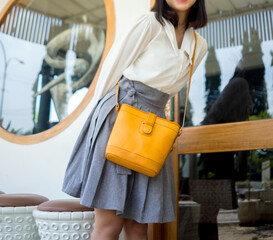 The body of a girl in a gray skirt and white shirt carrying a turmeric yellow leather bag, standing in front of a glass window.