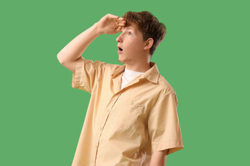 Surprised young man looking far away with hand over head on green background