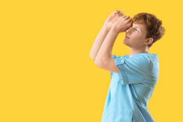 Young male doctor looking through imaginary binoculars on yellow background