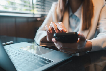 Professional woman multitasking with smartphone and laptop in a bright, modern office environment.