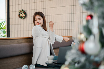 A young woman in a modern cafe stretches while working on her laptop, with festive Christmas decorations nearby.