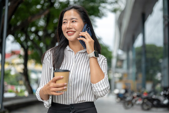 Asian woman office worker holding coffee cup as talking on phone while walking outside shopping mall