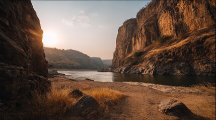 Bright sun sets behind tall rocky cliffs that frame a calm river. The warm light reflects off the water while dry grass and stones line the sandy shore in the fading day.