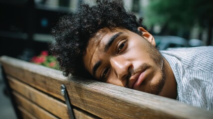 A young man with curly hair rests his head on a wooden bench in a city park. Flowers and green trees surround him as he enjoys a quiet moment on a sunny day.