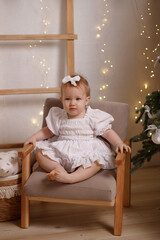 Caucasian baby girl, about 1 year old, in festive white dress with bow, smiling and waving on chair, front view, Christmas interior with lights. Concepts: holidays, joy, childhood, celebration. 