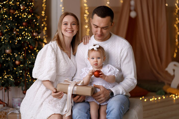 Young Caucasian couple and baby girl in white clothes, sitting together with gift box near Christmas tree, indoor, front view. Concept: holidays, love, parenthood, family bonding.