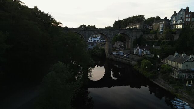 Drone flys around waterfront trees next to the river nidd to reveal a large stone built viaduct bridge with arc supports at sunset in Knaresborough