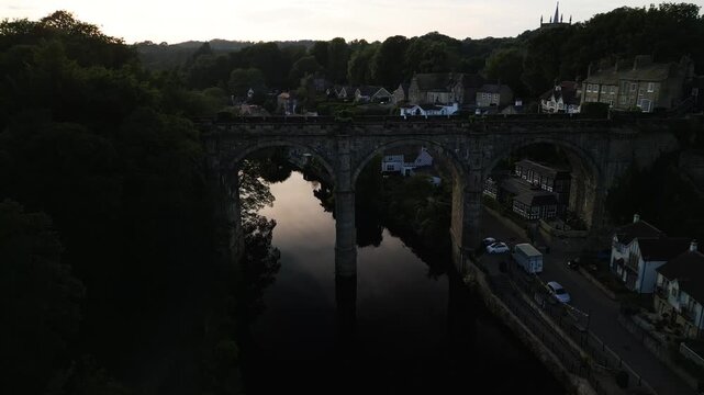 Drone flys above waterfront trees next to the river nidd to reveal a large stone built viaduct bridge with arc supports at sunset in Knaresborough