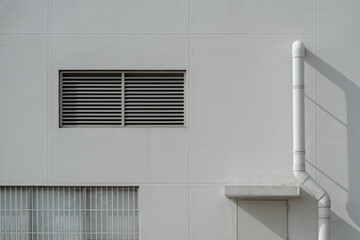 Industrial white wall facade featuring a louvered vent, barred window, and a vertical drainage pipe.
