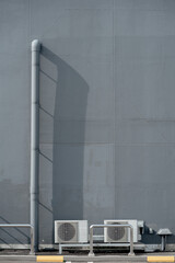 Two air conditioner outdoor units and a long drainage pipe on a gray textured industrial building wall.