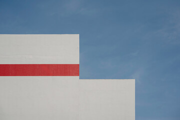 Minimalist corner of a white building with a red horizontal stripe against a clear blue sky.