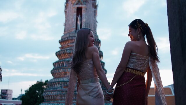 Thai women holding hands, smiling at wat arun temple, wearing chut thai