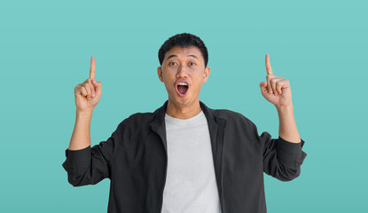 A young Asian man wearing black coat happy shocked face pointing thumbs up isolated on aqua background.